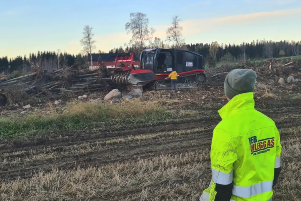 En mann i gul jakke står foran en haug med tømmerstokker i en park.