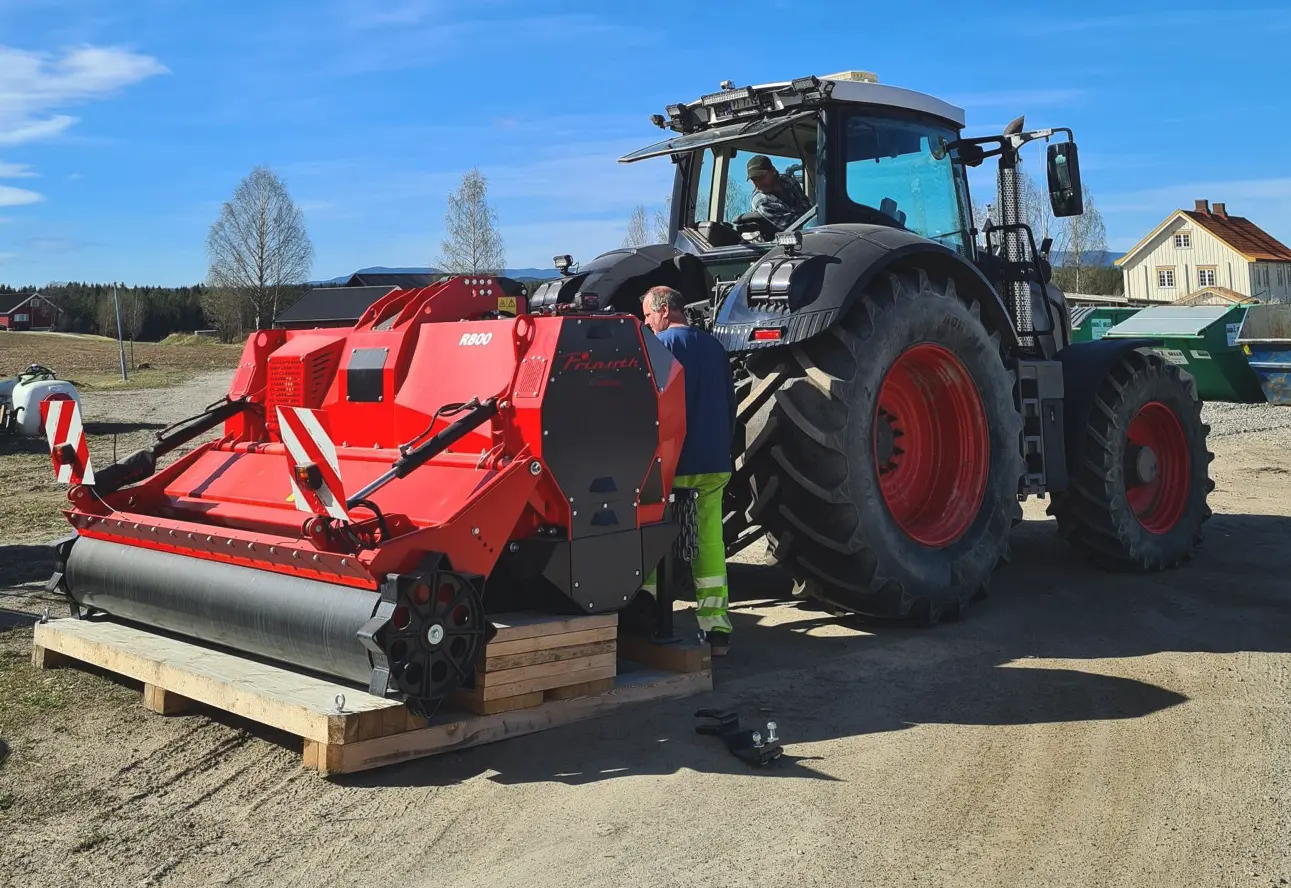 En mann står ved siden av en rød traktor på en grusvei. Mannen er ansatt i selskapet med ansvar for vedlikehold og drift av traktorene. Han utstråler pålitelighet